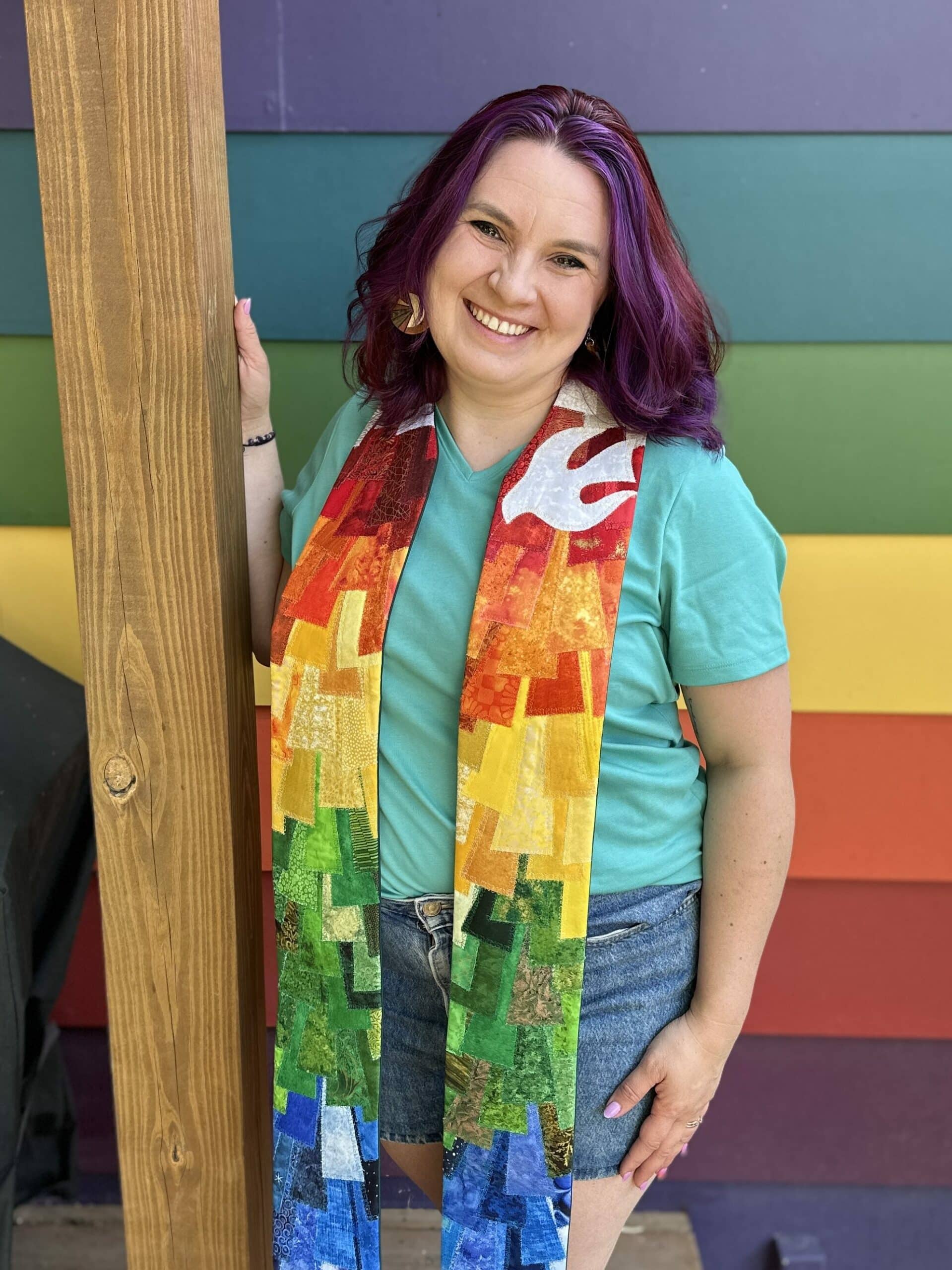 a woman with purple hair is wearing a rainbow stole and smiles at the camera in front of a rainbow wall